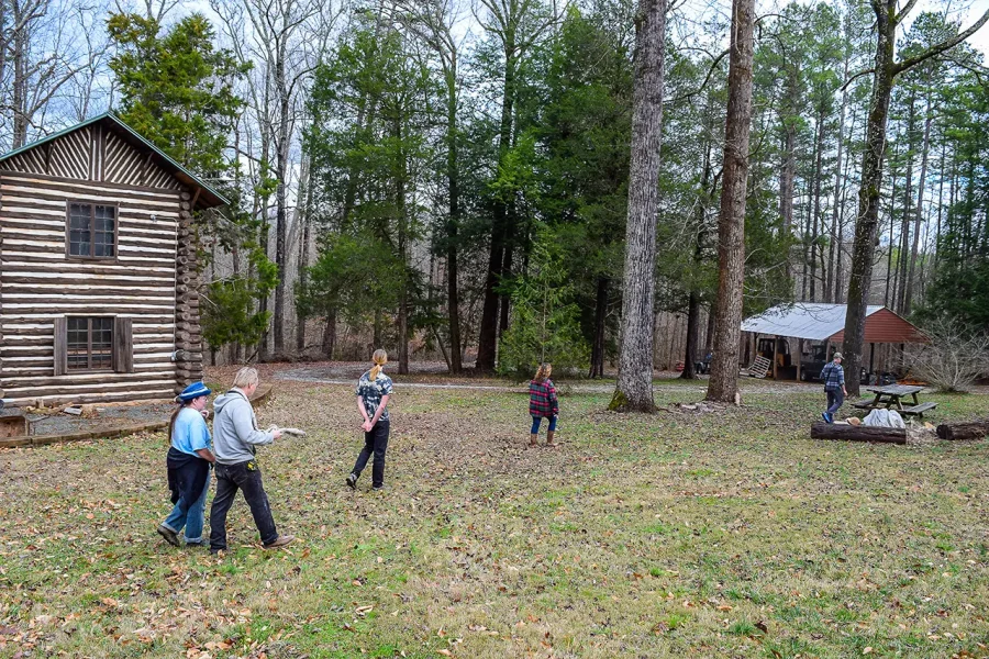 people walking across a clearing surrounded by trees and a log cabin